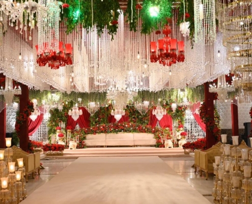 A lavish wedding stage with cascading crystal chandeliers, red and green floral arrangements, and an elegant white aisle leading to a grand seating area.